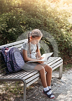 Girl with a backpack sitting on a bench and reading a book near the school. Back to school, lesson schedule, a diary with grades.