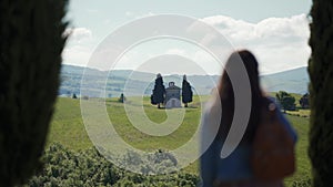 Girl with backpack stands, enjoys scenic Tuscan landscape with Vitaleta chapel