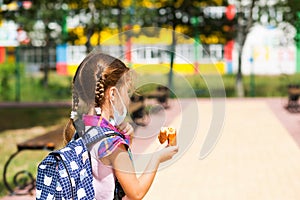 Girl with a backpack take off the medical mask and eating pie near the school. A quick snack with a bun, unhealthy food, lunch