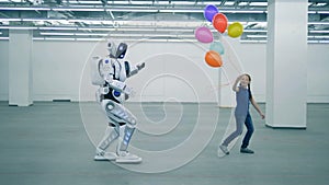 A girl with balloons is dancing and a manlike robot is coming towards her