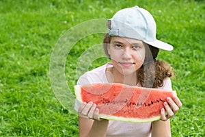 A girl in a baseball cap holds a large slice of watermelon in her hands in front of her