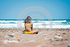 Girl in a bathing suit is sitting on a sandy beach watching the waves and the sea