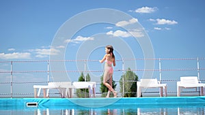 Girl in bathing suit stands between sun loungers by pool background sky