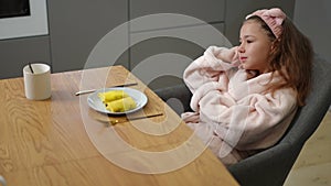 A girl in a bathrobe and a hair band on her head sits on a chair and dreams at the table, on which there is a plate with