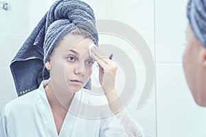 The girl in the bathroom in front of the mirror with a towel on her head applies cream to her face. selective focus. film grain