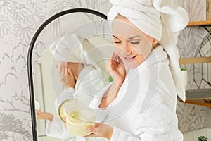 A girl in the bathroom with a towel on her head and in a bathrobe holds a face cream in her hand