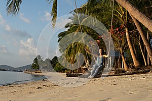 The girl on the beach rides on a swing during sunset. Sunset in the tropics, enjoying nature. Swing tied to a palm tree by the