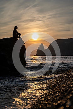 A girl on the beach watches the sunset on the sea. She's sitting on a rock in the sea. The sun is setting behind the