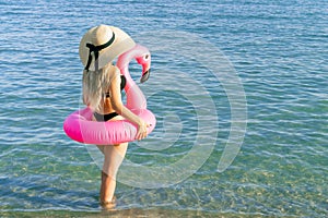 Girl by beach. Young sexy woman in straw hat, bikini swimsuit, sunglasses with pink inflatable flamingo in blue ocean water for