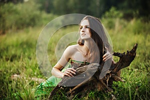 Girl in a beautiful green dress lies on the grass near the roots of a tree and dreams. Woman with bright makeup in nature, natural