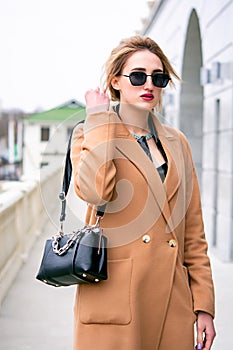 A girl in a beige coat, black glasses, a black bag in her hands