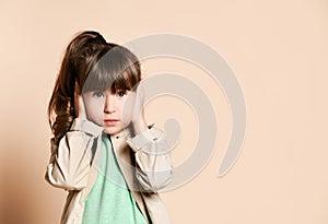 A girl in a beige coat stands with her hands over her ears.