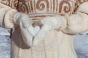 A girl in a beige sweater holds a heart made of snow in her hands, dressed in white mittens, from the side.