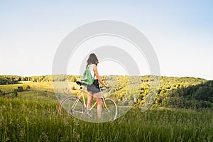 Girl with bicycle enjoying beautiful rural landscape. Young pretty female person with retro bike standing in a meadow on bright s