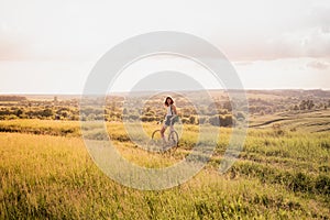 Girl with a bicycle posing on the hill in beautiful rural landscape at sunset. Young pretty female person with retro bike