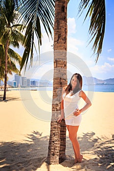 girl with big bust in white frock poses by palm on beach