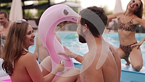 Girl in bikini hanging out with boyfriend on pool party. Couple kisses having fun on inflatable pink flamingo float