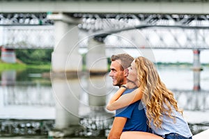 The girl bites an ear to the guy. Happy romantic caucasian couple looking at a lake