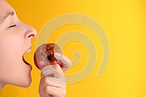 The girl bites a rotten apple with a worm on a yellow background. Expired products, junk food