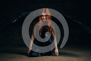 A girl with black angel wings sits on the floor in a dark room. Red-haired girl