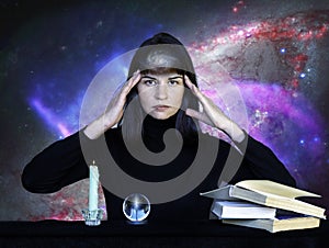 A girl in black clothes sitting at a table with books, a sphere and a burning candle against background of starry sky