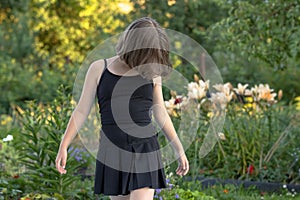 A girl in black clothes stands with her head bowed and her face covered with long hair in a blooming summer garden