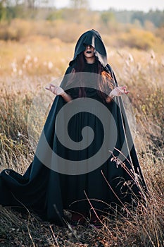 A girl in a black dress, a cloak with a hood stands in a high dry grass in the field against the background of the forest. Witch C