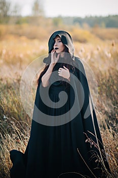 A girl in a black dress, a cloak with a hood stands in a high dry grass in the field against the background of the forest. Witch C