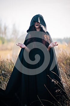 A girl in a black dress, a cloak with a hood stands in a high dry grass in the field against the background of the forest. Witch C