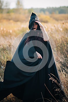 A girl in a black dress, a cloak with a hood stands in a high dry grass in the field against the background of the forest. Witch C photo