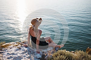 The girl in a black dress and hat on a rocky beach looks at the sea