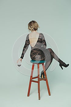 A girl in a black guipure dress sits with her back on a bar stool holding a glass of wine in her hand