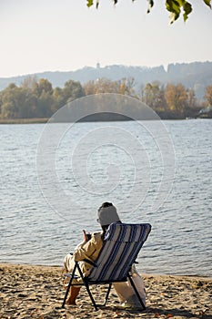 A girl with black hair sits on a riverbank in a deck chair and looks into a mobile phone