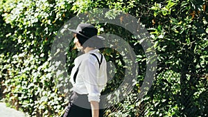 A girl in a black hat and suspenders goes along of green bushes at beautiful day