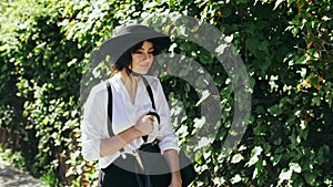 A girl in a black hat and suspenders goes along of green bushes at beautiful day