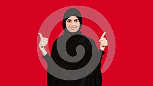 Girl in black hijab smiles and poses at the camera showing two arms like. Thumbs up on a red background with side space.