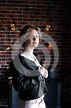 The girl in a black jacket and a light blouse poses opposite to a window, a background the brick wall covered with glow lamps.