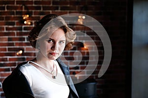 The girl in a black jacket and a light blouse poses opposite to a window, a background the brick wall covered with glow lamps.