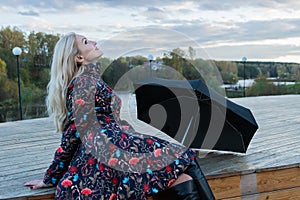 A girl with a black umbrella sits on a wooden staircase, a lot of puffy clouds on the stage. with charismatic appearance