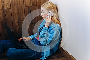 Girl with blond hair in denim clothes working at home with a laptop and talking on the phone.