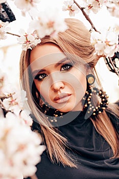 Girl with blond hair in elegant clothes and earrings posing among flowering peach trees