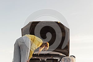 a girl with blond hair in a yellow sweater and jeans, stands with the hood of the car open and looks at what has broken