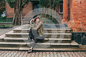 Girl with blond long hair, in black leggings, a tank top, a beige jacket and a hat, sits on the stairs