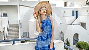Girl blonde with long hair in a straw hat in a blue dress with polka dots stands in Santorini