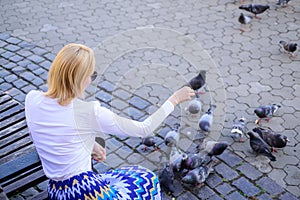 Girl blonde woman relaxing city square and feeding pigeons. Woman tourist or citizen toss crumbs for pigeons. Girl