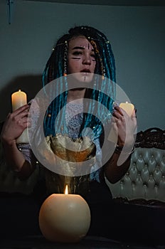 Girl with blue dreadlocks conjures at night in a dark room, candle fire and a cow skull .