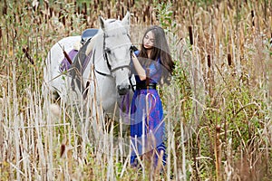 Girl in blue dress holds by bridle white horse