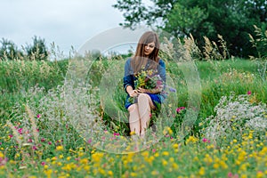 A girl in a blue dress and jean jacket with a bouquet of wildflowers is sitting between a lot of flowers.