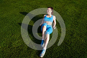 Girl in blue fitness clothes lying on grass stadium. Brunette advertises green lawn on football field. Woman has sporty lifestyle