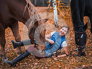 A girl in a blue jacket lies next to the horse and laughs. Golden autumn background